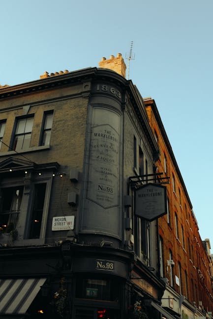 Photograph of a historic corner building on Marylebone High Street, with a rounded facade and large windows. The building has a light grey stone exterior with decorative architectural details at the roofline. A vintage-style sign hangs on the side, displaying the name 'The Marylebone' along with other faded text, indicating its historical significance. The ground floor features a storefront with a striped awning, and a small street sign indicating 'Marylebone Street W1' is visible. The building is illuminated softly by natural daylight, highlighting its classic design and well-maintained appearance. This image is relevant to the context of best cleaning tips for Marylebone High Street flats, as it depicts an iconic local structure, emphasizing the importance of maintaining historical and residential properties through expert cleaning services. Marylebone Cleaners specializes in comprehensive domestic and commercial cleaning, ensuring buildings like this remain pristine and well-preserved.