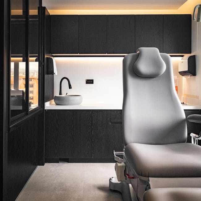 A modern medical examination room featuring a clean, white examination chair with a smooth, leather-like surface, positioned beside a sleek, white countertop with a contemporary round ceramic basin and a black faucet. The background includes dark matte cabinetry with minimalistic design, mounted above the countertop, and a white wall with an electrical outlet. The room is well-lit with ambient lighting providing a warm atmosphere, and the floor is covered in light-colored tiles, all appearing spotless and ready for sanitisation. Visible cleaning tools or supplies are not in the scene, but the overall environment reflects professional surface cleaning and hygiene standards, consistent with the services provided by Marylebone Cleaners for healthcare clinic settings.