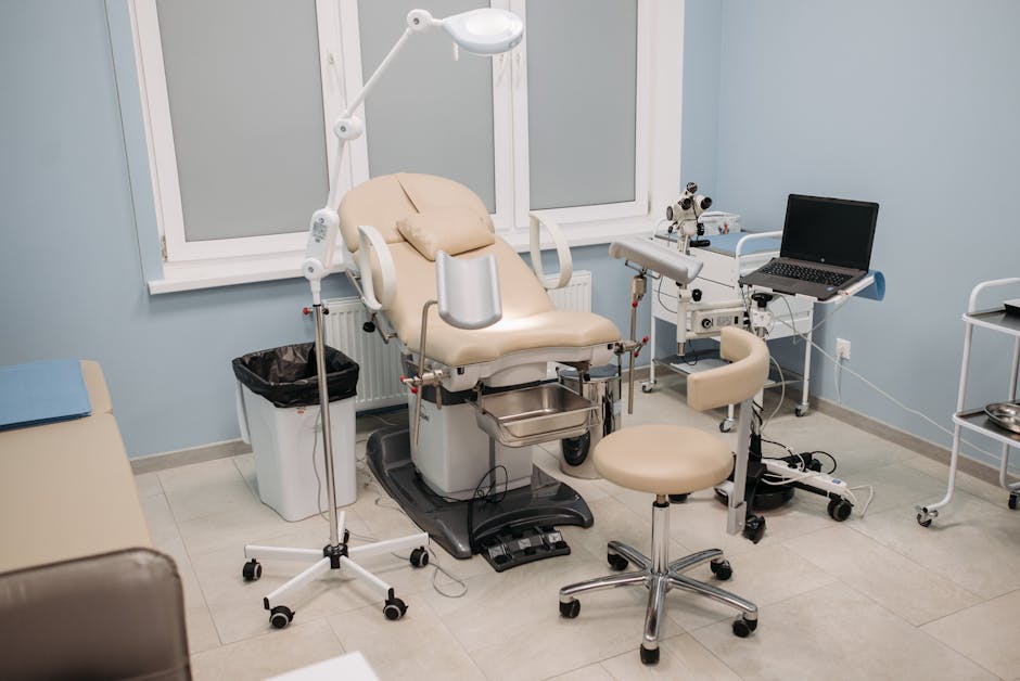 A medical examination room featuring a beige reclining examination chair with an adjustable headrest and armrests, positioned in front of a light blue wall with three large windows. The room has a white floor and is equipped with various medical tools, including a white overhead lamp on a stand, a small stainless steel tray, a laptop on a mobile cart, and a microscope mounted on a white stand. A small black waste bin is placed next to the examination chair, and a cleaning cloth is visible on an adjacent chair. The room appears clean and hygienic, with bright natural lighting coming through the windows, and organized equipment facilitating surface cleaning and sanitisation, as provided by Marylebone Cleaners, specialists in deep cleaning and hygiene maintenance for clinical environments.