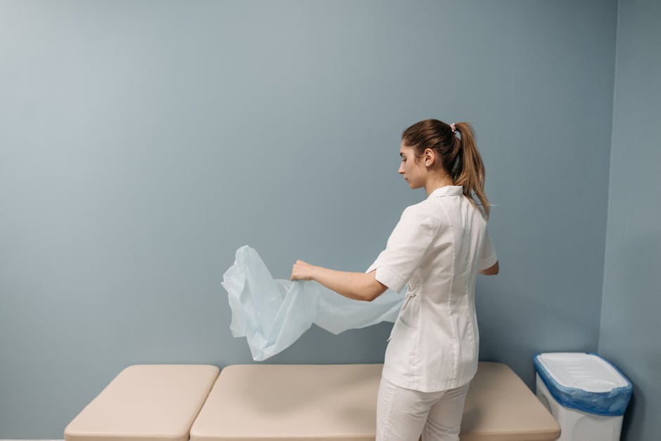A modern medical examination room featuring a clean, white examination chair with a smooth, leather-like surface, positioned beside a sleek, white countertop with a contemporary round ceramic basin and a black faucet. The background includes dark matte cabinetry with minimalistic design, mounted above the countertop, and a white wall with an electrical outlet. The room is well-lit with ambient lighting providing a warm atmosphere, and the floor is covered in light-colored tiles, all appearing spotless and ready for sanitisation. Visible cleaning tools or supplies are not in the scene, but the overall environment reflects professional surface cleaning and hygiene standards, consistent with the services provided by Marylebone Cleaners for healthcare clinic settings.