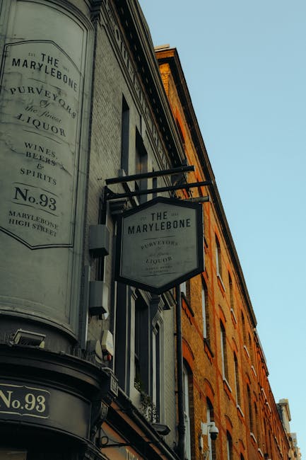 The image depicts the exterior of a historic building with a curved facade, showcasing signage for The Marylebone pub, which includes a vintage-style signboard mounted with black metal brackets and a white background. The building features large windows with black frames and stone trim, and a combination of grey and brick-colored surfaces. The scene is illuminated by natural daylight, highlighting the building's clean and well-maintained appearance, consistent with professional surface cleaning and maintenance standards associated with domestic or commercial cleaning services, such as those provided by Marylebone Cleaners, especially relevant for ensuring the cleanliness of building exteriors in the context of permits for skip hire in Marylebone W1.