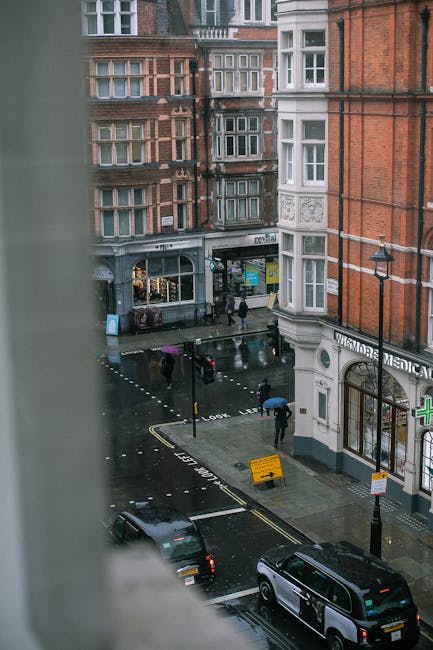View of a rainy city street in Marylebone, showing wet asphalt pavement with reflections of surrounding buildings and streetlights. Several pedestrians with umbrellas walk along the sidewalk near storefronts, including a medical clinic with a green cross sign and a retail shop with display windows. Parked cars line the street, and a streetlamp stands on the corner. The image captures the exterior urban environment, emphasizing cleanliness maintenance of the outdoor surfaces in a typical residential and commercial area. For interior surface cleaning and sanitisation, Marylebone Cleaners offers professional domestic and commercial cleaning services tailored to properties affected by tenancy disputes, ensuring a thorough and hygienic refresh after occupancy changes.