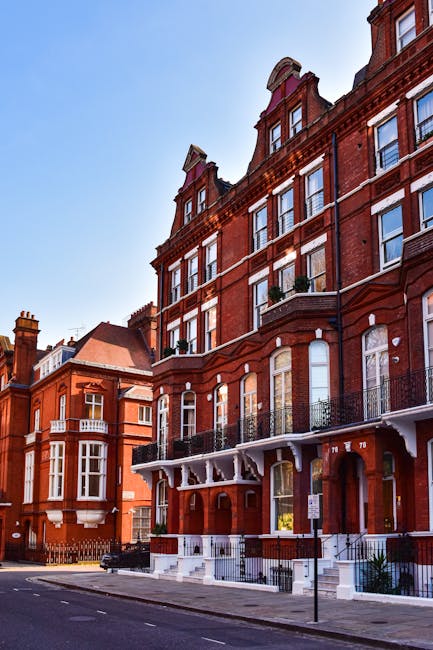 A street view of a historic red brick building in Marylebone, featuring multiple floors with large sash windows and decorative architraves, illuminated by natural daylight. The building has black wrought iron balconies on the upper levels, with some potted plants visible, and a ground-floor entrance with stairs leading up to a glass door. The sidewalk in front is clean and free of debris, with a small street sign and parked cars nearby, reflecting a well-maintained urban residential area. This scene is related to domestic cleaning and property maintenance, emphasizing cleanliness and hygiene, consistent with services provided by Marylebone Cleaners for emergency flat cleans after tenancy disputes.