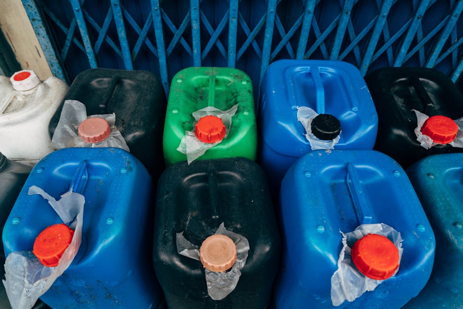 A collection of nine plastic containers with various colored caps, including white, black, red, and green, arranged on a surface against a blue metal fence. The containers appear to be used for storing liquids and are somewhat dusty, with some showing signs of wear and tear. The surface is partially visible, and the containers are positioned in close proximity to each other, with some of the caps covered with torn bits of paper or plastic. The image demonstrates a variety of materials including durable plastic, and the containers are placed outdoors under natural lighting conditions, suggesting the need for proper cleaning and storage for safety and compliance with hazardous waste removal laws. This scene reflects the importance of surface cleaning and hygienic maintenance that Marylebone Cleaners can provide, especially in contexts involving hazardous waste management.
