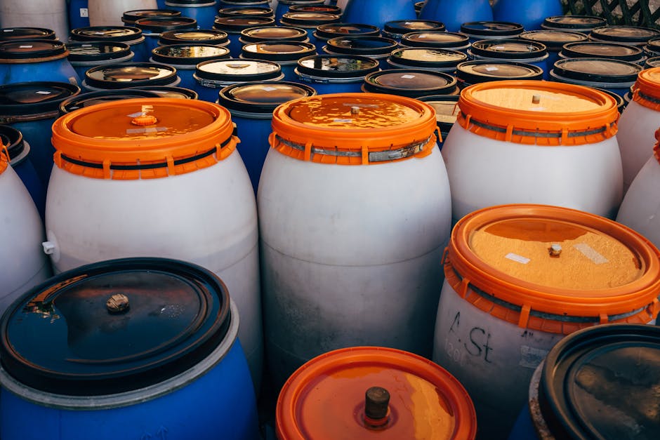 A collection of large plastic drums and barrels, predominantly white with orange and black lids, arranged outdoors on a surface. The drums vary in height and some have visible labels or markings. They are positioned in a tight cluster, with the focus on the foreground barrels, which are clean and sealed, while the background barrels appear slightly blurred. The lighting suggests natural daylight, highlighting the glossy surfaces of the drums and the reflective qualities of the sealed lids. This setup is indicative of hazardous waste storage, relevant to hazardous waste removal laws for Marylebone landlords. For professional surface cleaning, sanitisation, or deep cleaning services, Marylebone Cleaners offers expert assistance to ensure safety and compliance.