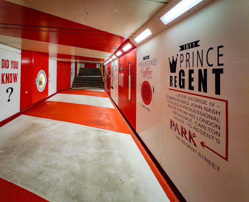 An interior corridor featuring a polished concrete floor with red and white painted sections, illuminated by bright white ceiling lights. The walls are decorated with historical and promotional signage in black and red text, including the name 'Prince Regent' with a crown motif, and directional arrows. A staircase with dark wooden steps and metal railings is visible at the end of the corridor. The space is clean and appears well-maintained, with no dust or dirt evident on surfaces. This setting exemplifies surface cleaning and maintenance standards on behalf of Marylebone Cleaners, demonstrating their expertise in ensuring hygienic, spotless environments in commercial or residential buildings.