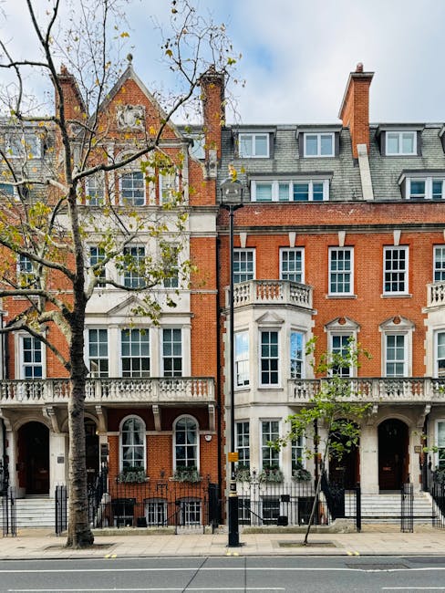 A view of a brick residential building on a street in Marylebone, London, with white-framed double-hung sash windows and street signs indicating Balderton Street W1 and Providence Court W1. The scene includes a narrow alleyway with black wheelie bins placed alongside the building, a black wrought iron fence at the lower part of the image, and two people walking in the background. The lighting suggests daytime with natural light illuminating the scene. The image reflects the urban setting typical of Marylebone, showcasing red brick facades and street signage, with no visible cleaning activity. The scene aligns with the work of Marylebone Cleaners in maintaining residential environments, which often involves surface cleaning, deep cleaning, and sanitisation in similar urban settings.
