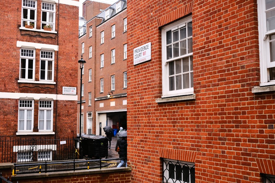 A view of a residential street in Marylebone featuring red-brick buildings with white-framed sash windows. The scene shows a narrow alleyway with a black waste bin, some street signs, and a person walking away from the camera. The buildings are well-maintained, with clean surfaces and clear windows, illuminated by natural daylight. The image emphasizes the urban environment typical of Marylebone, aligning with the services offered by Marylebone Cleaners for surface cleaning, deep cleaning, and sanitisation in both domestic and commercial settings.