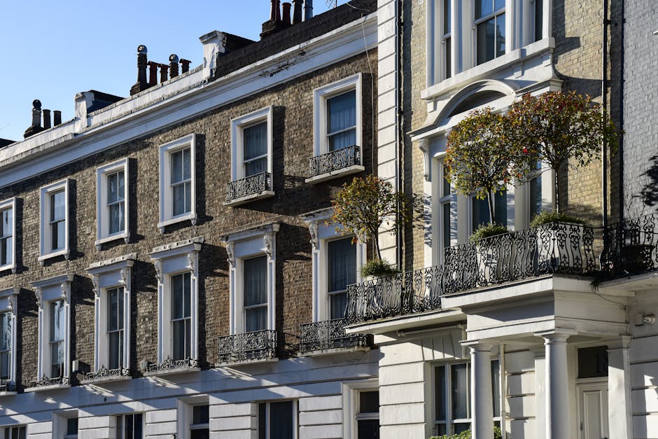 Photograph of a row of classic London townhouses with brick facades, featuring multiple white-framed sash windows and ornate black wrought iron balcony railings. Some balconies have potted plants and small trees, contributing to a clean and well-maintained appearance. The ground level shows a white-painted section with neatly aligned windows, and the buildings are illuminated by bright sunlight, casting shadows on the surfaces. The overall scene emphasizes the exterior architectural details and cleanliness of residential buildings typical in Marylebone, aligning with professional deep cleaning and surface sanitisation standards promoted by Marylebone Cleaners in the W1 postcode area.