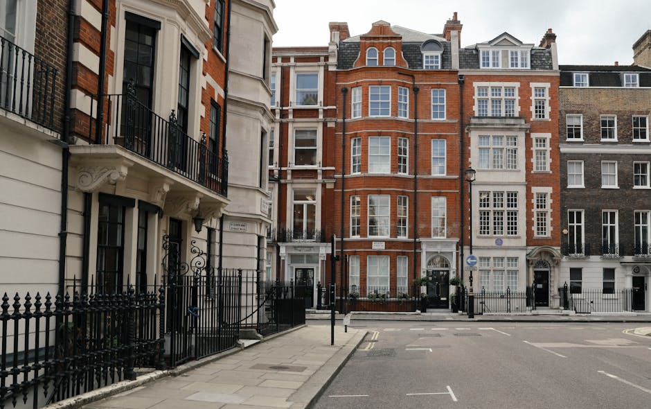 Photograph of a typical residential street in Marylebone, London, featuring elegant multi-storey Victorian and Edwardian houses with brick and stucco facades, large sash windows, and decorative architectural details. The street is paved with grey asphalt, marked with parking lines, and lined with black wrought iron fences and traditional lampposts. The buildings are well-maintained, with clean exteriors and tidy entrances, reflecting high standards of property upkeep associated with the W1 postcode. The scene is illuminated by natural daylight, emphasizing the neat and pristine condition of the surroundings, consistent with the high cleaning standards promoted by Marylebone Cleaners for domestic and commercial properties.