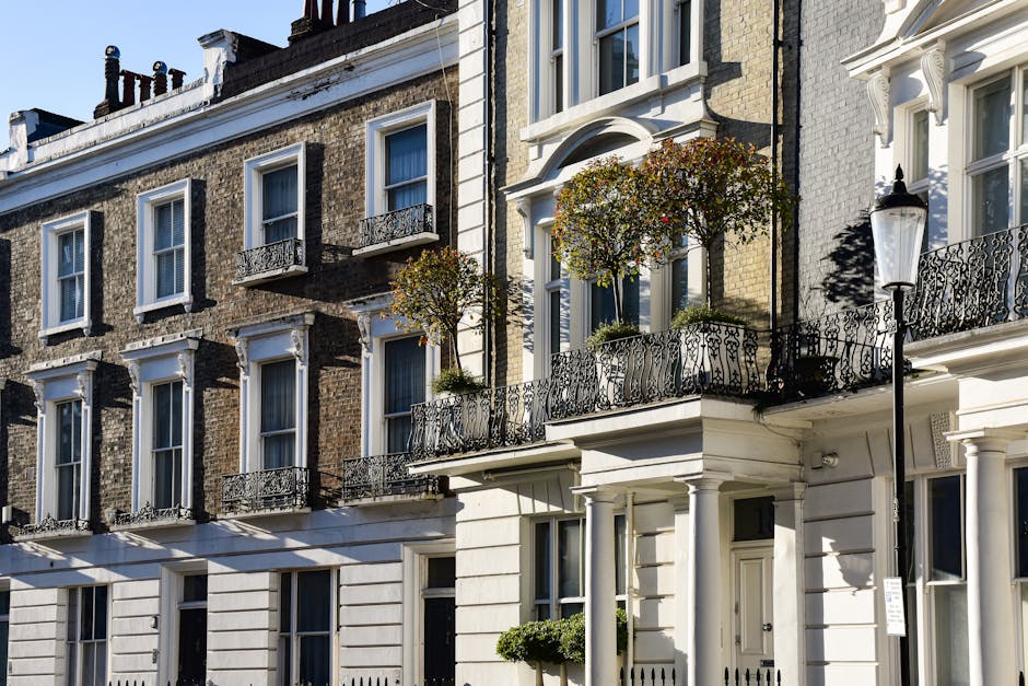Photograph of a row of classic London townhouses with brick facades, featuring multiple white-framed sash windows and ornate black wrought iron balcony railings. Some balconies have potted plants and small trees, contributing to a clean and well-maintained appearance. The ground level shows a white-painted section with neatly aligned windows, and the buildings are illuminated by bright sunlight, casting shadows on the surfaces. The overall scene emphasizes the exterior architectural details and cleanliness of residential buildings typical in Marylebone, aligning with professional deep cleaning and surface sanitisation standards promoted by Marylebone Cleaners in the W1 postcode area.