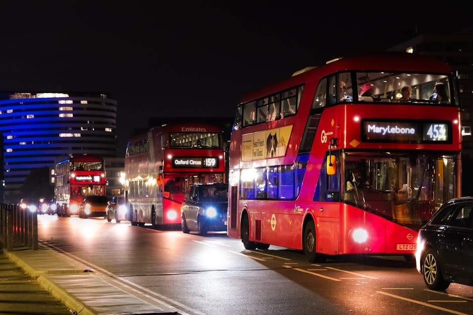 A lineup of red double-decker buses parked along a city street at night, illuminated by streetlights and vehicle headlights. The bus in the foreground displays the route number 453 heading to Marylebone, while others behind show route 159 and Emirates branding. The street features a smooth asphalt surface, with clearly marked lanes and some parked cars beside the buses. Modern buildings with illuminated windows are visible in the background, casting a soft glow against the dark sky. The scene emphasizes urban public transportation, cleanliness, and the organized layout of a bustling city street, consistent with the principles of surface cleaning and maintenance as outlined in Westminster Council rules on waste disposal for Marylebone, managed by Marylebone Cleaners.