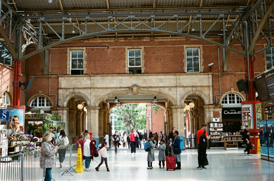The image shows a spacious indoor area of a train or bus station with high ceilings and brick archways, illuminated by natural light through large windows and complemented by hanging pendant lights. The floor is smooth, polished, and clean, reflecting the ambient lighting. In the foreground, there are people walking, some holding luggage or backpacks, with a few engaged in conversations or checking their phones. To the left, a flower stand displays vibrant bouquets and potted plants, grouped neatly on metal shelves. Nearby, a yellow caution sign is placed on the floor, indicating a recent or ongoing cleaning activity. On the right, a small retail kiosk offers chocolates and travel essentials, with a few customers browsing. The entire setting exudes a tidy, well-maintained atmosphere consistent with professional interior cleaning and surface sanitisation standards. Marylebone Cleaners provides expert domestic and commercial cleaning services, supporting cleanliness and hygiene in busy public spaces such as this, especially after clearance or movement of bulky waste materials. The environment highlights the importance of surface cleaning, deep cleaning, and sanitisation to maintain a safe and welcoming space for travelers and visitors.
