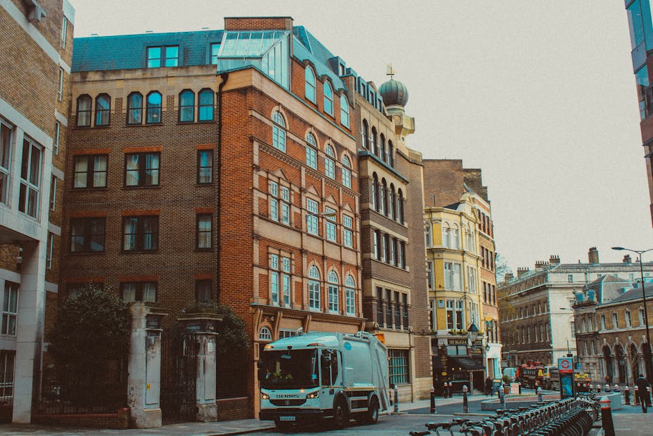 A street scene in Marylebone featuring a row of multi-storey buildings with a mix of brick, stone, and ornate architectural details. The buildings display large windows, some with decorative arches, and a variety of roof styles including a rounded dome. In the foreground, a street cleaning vehicle, likely used for deep cleaning and surface sanitisation, is parked alongside a paved road with metal bike racks and bollards. The area appears clean and well-maintained, with minimal litter visible on the street. Surrounding the buildings are street lamp posts, signage, and a few trees, all under natural daylight, highlighting the urban environment typical of Marylebone. This image subtly reflects the importance of maintaining cleanliness in busy city districts, which [COMPANY_NAME] offers through comprehensive domestic cleaning and surface sanitation services, especially after clearance or bulky waste removal.